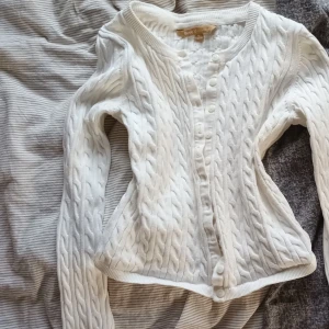 Vit kabelstickad kofta Basic U, S - Supersöt vit kabelstickad kofta från Basic U collection i storlek S. Koftan har långa ärmar, rund halsringning och knäppning framtill med vita knappar. Perfekt för lager-på-lager och ger en clean och klassisk look.