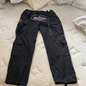 Svarta cargopants från Corteiz S - Svarta cargopants från Corteiz med flera fickor, broderad logga och lila patchdetaljer. Byxorna har bälte i midjan och coola broderier på benet. Perfekta för dig som gillar streetwear och vill sticka ut med snygga detaljer.