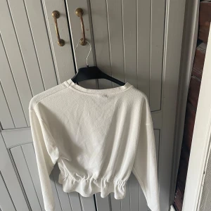 Vit långärmad tröja med snörning - Säljer en vit långärmad tröja med rund hals och snygg snörning i midjan som ger en croppad och avslappnad look. Tröjan har en lätt struktur i tyget och passar perfekt till jeans eller kjol.