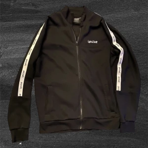 Svart ziptröja Lyle & Scott S - Svart ziptröja från Lyle & Scott med vita band längs ärmarna där loggan är tryckt. Tröjan har hög krage, hel dragkedja och två fickor framtill. Perfekt för en sportig och clean look.