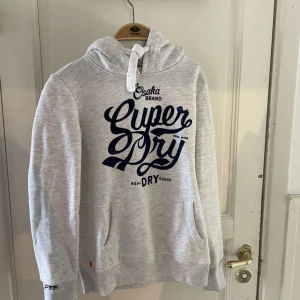 Ljusgrå hoodie från Superdry - Säljer en ljusgrå hoodie från Superdry med stor mörkblå logga på bröstet. Perfekt för dig som gillar streetstyle och vill ha en skön och snygg tröja till vardags.