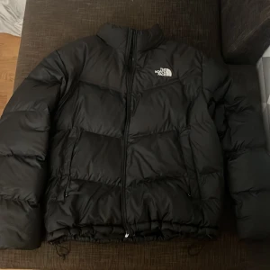 Svart pufferjacka från The North Face - Säljer en svart pufferjacka från The North Face i storlek L då efter jag beställt den online så var den en storlek för stor för mig. Användes endast två gånger förra vintern så den är nästan helt ny. Hög krage och två fickor gör den simpel men fräsch. 