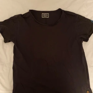 Svart t-shirt från Replay - Svart t-shirt från Replay med klassisk passform och rund hals. T-shirten har tryckt Replay-logga i blått på ena sidan och en gul Replay-patch på ärmen. Tillverkad i mjuk bomull som är skön mot huden. Perfekt för en chill och stilren look.