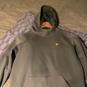 Grön hoodie från Lyle & Scott. Passar M och S - Snygg mörkgrön hoodie från Lyle & Scott med klassisk gul fågel-logga på bröstet. Tröjan har huva med snörning, magficka och ribbade muddar vid ärmslut och nederkant. Perfekt för chill dagar och enkel att matcha med jeans eller joggers.