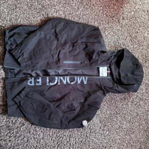 Svart Moncler vindjacka med luva - Svart vindjacka från Moncler med stor logga på bröstet och klassisk patch på ärmen. Jackan har huva med dragsko, hel dragkedja framtill och praktiska fickor med zip. Materialet är lätt och syntetiskt, perfekt för blåsiga dagar. Snygg och stilren design med Moncler-detaljer.