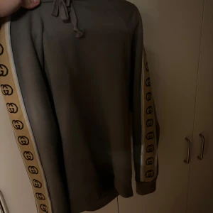 Gucci hoodie med logoband på ärmarna - Säljer en snygg mörkgrå hoodie från Gucci med klassiska logoband i beige och svart längs ärmarna. Tröjan har huva med snörning och en relaxed passform. Perfekt för dig som vill sticka ut med exklusiv streetstyle.