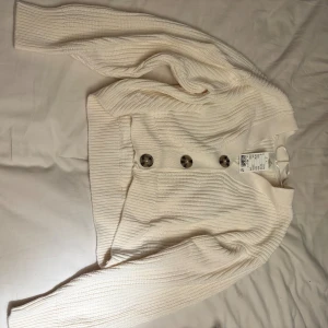 Ribbad vit kofta med stora knappar - Säljer en ribbad vit kofta från H&M med stora, mönstrade knappar framtill. Koftan har lång ärm och är gjord i mjukt stickat material som känns skönt mot huden. Perfekt att slänga på sig när det är lite kyligt ute.