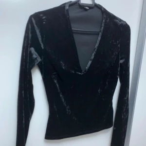Svart sammetstopp med djup v-ringning - Säljer en svart långärmad topp i mjuk sammet med djup v-ringning. Toppen har en figurnära passform och är perfekt för dig som gillar en stilren men ändå lyxig look. Materialet ger en snygg glans och känns riktigt skönt mot huden.