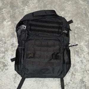 Svart taktisk ryggsäck med spännen HELT NY - En svart ryggsäck i slitstark syntet med flera fack, justerbara remmar och praktiska spännen framtill. Utrustad med meshfickor på sidorna och MOLLE-system för extra utrustning. Perfekt för dig som gillar funktionell och stilren design. HELT NY