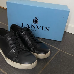Svarta Lanvin sneakers i skinn - Snygga svarta sneakers från Lanvin med rund tå och platt vit sula. Skorna är tillverkade i mjukt skinn och har klassisk snörning med metallöglor. Perfekt för dig som gillar stilrena och exklusiva sneakers med en lyxig känsla.