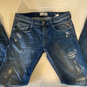 Blå Replay jeans med slitningar - Säljer ett par blå jeans från Replay med snygga slitningar och tvättade detaljer. Klassisk femficksmodell med logga på bakfickan och läderpatch i midjan. Jeansen har normal passform och är tillverkade i bomull för en skön känsla.