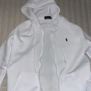 Vit hoodie från Polo Ralph Lauren - Vit hoodie från Polo Ralph Lauren med klassisk huva och snörning. Tröjan har en broderad logga på bröstet och känguruficka framtill. Perfekt för en clean och stilren look.