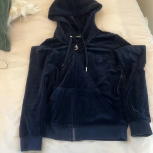 Mörkblå velour hoodie Juicy Couture XS - Mörkblå hoodie från Juicy Couture i mjuk velour med huva och snörning. Klassisk dragkedja framtill och två fickor. Perfekt för en chill och trendig look.
