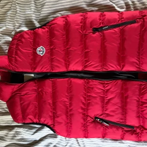 Röd dunväst från Moncler - Säljer en röd dunväst från Moncler med klassisk logga på bröstet. Västen har svarta dragkedjor framtill och på fickorna, quiltad design och en cool serietidningsetikett på insidan. Perfekt för lager-på-lager och streetstyle.  Pris kan diskuteras fraktar snabbt 