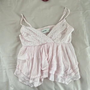 Ljusrosa spetslinne från Hollister - Supersött ljusrosa linne från Hollister med tunna axelband och v-ringning. Överdelen har fina spetsdetaljer och nedtill är tyget luftigt och lätt rynkat för en flowy känsla. Perfekt för varma dagar och somriga vibes!