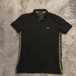 Svart pikétröja Hugo Boss slim fit - Svart pikétröja från Hugo Boss med slim fit-passform. Tröjan har guldiga detaljer och logga längs sidorna samt broderad Boss-logga på bröstet. Klassisk krage och korta ärmar. Perfekt för dig som gillar stilrena plagg.