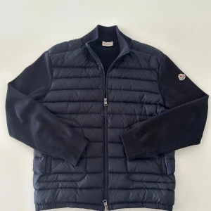 Moncler cardigan  - Säljer en marinblå cardigan från Moncler med stickade ärmar och quiltad front. Jackan har dragkedja, ribbade muddar och Moncler-logga på ena ärmen. Fylld med vitt gåsdun för extra värme. Perfekt för dig som vill ha en clean och stilren look. Hör av er vid frågor