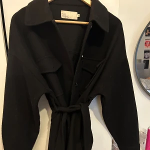 Svart kappa från NLY Trend med bälte - Svart kappa från NLY Trend i storlek 36. Jackan har stora bröstfickor med lock, knappar hela vägen och ett matchande bälte i midjan. Rak passform med klassisk krage och långa ärmar. Perfekt för lager-på-lager och snygg till jeans eller dressade byxor.