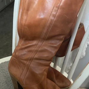 Snygga bruna skinnstövlar med klack - Snygga bruna stövlar i äkta skinn med rund tå och stabil klack. Stövlarna har en klassisk design med dekorativa sömmar längs skaftet och en träfärgad klack som ger extra höjd. Perfekta för dig som gillar en tidlös och cool look. Stl:40