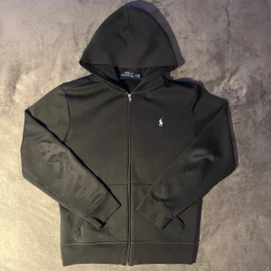 Svart zip hoodie från Polo Ralph Lauren - Svart hoodie från Polo Ralph Lauren med dragkedja och huva. Tröjan har det ikoniska broderade logotypen på bröstet och är tillverkad i mjuk bomull. Tröja är i ett mycket bra skick och det finns inga deflekter. Tröjan är i storlek M.