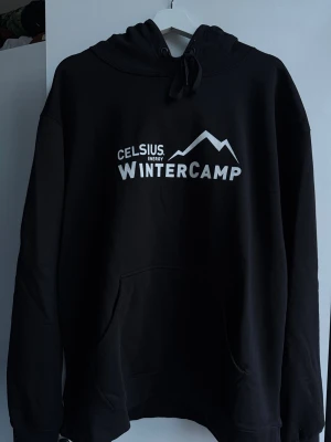 Svart Celsius Wintercamp hoodie - Svart hoodie från Celsius med vit text 'WINTERCAMP' och bergmotiv på bröstet. Klassisk känguruficka framtill, justerbar huva med snören och mjukt material som känns skönt mot huden. Perfekt för dig som gillar streetwear och vill ha en chill look.