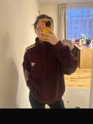 Vinröd Vintage y2k Adidas hoodie med dragkedja - Säljer en vinröd hoodie från Adidas med dragkedja och klassiska vita ränder längs ärmarna. Tröjan har hög krage, två fickor och Adidas logga på bröstet. Perfekt för en sportig och avslappnad stil.