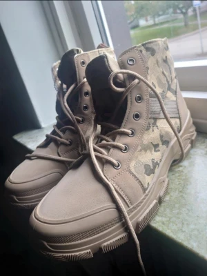 Beige kängor med camomönster - Säljer ett par beige kängor med camouflagemönster på sidorna. De har snörning framtill, rund tå och platt sula. Materialet är en mix av bomull och syntet, vilket gör dem både lätta och slitstarka. Perfekta för dig som gillar streetwear och vill sticka ut.