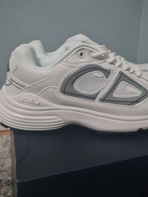 Vita Dior B30 sneakers - Säljer ett par riktigt snygga Dior B30 sneakers i vitt med gråa detaljer och stor CD-logga på sidan. Skorna har meshpaneler för extra andningsförmåga och en chunky sula som ger grym streetstyle-vibe. Klassisk snörning och exklusiv känsla, perfekta för dig som gillar lyxiga sneakers.