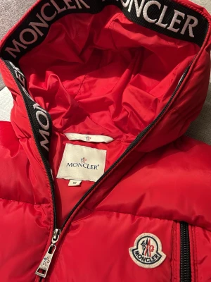 Röd dunväst ifrån moncler - Riktigt snygg röd dunväst från Moncler med huva och dragkedja framtill. Västen har två stora fickor med dragkedja och Moncler-logga på bröstet. Svarta detaljer med Moncler-tryck på huvkanten ger en exklusiv känsla. Västen är köpt ifrån butiken Vintage and friends.