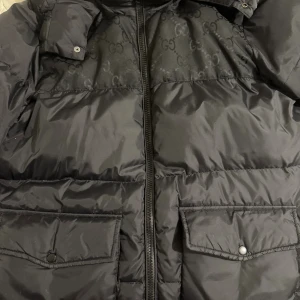 Svart pufferjacka från Gucci - Svart pufferjacka från Gucci med diskret GG-mönster upptill och stora fickor framtill. Jackan har dragkedja och klassisk pufferdesign, perfekt för kyliga dagar. Snygg och lyxig känsla med märkets ikoniska detaljer. Storlek 50 passar L/XL. Vid frågor kontakta mig! 