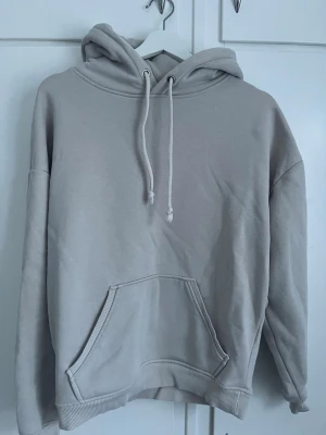 Beige hoodie med magficka - En stilren beige hoodie med stor magficka och justerbar huva med dragsko. Hoodien har ribbade muddar vid ärmslut och nederkant. Perfekt för en avslappnad och trendig look. Materialet känns mjukt och bekvämt.