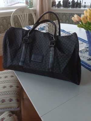 Duffelbag från Giorgio Armani - Snygg svart resväska från Giorgio Armani med diskret mönster och dubbla handtag. Väskan har ett klassiskt och stilrent utseende med logotyp framtill. Perfekt för weekendresor och har gott om plats för packning. Allt är i nyskick förutom att en läderbit har gått sönder, därav lågt pris