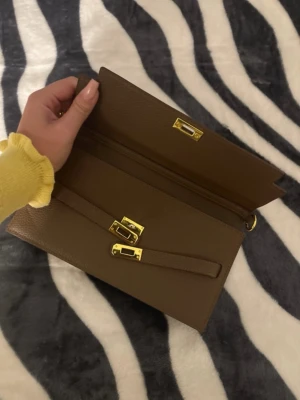 Brun kuvertväska med gulddetaljer - Snygg brun kuvertväska i skinn med guldfärgade spännen och dragkedja. Väskan har en stilren design med struktur i materialet och flera fack inuti för smart förvaring. Perfekt accessoar för att lyfta din outfit.