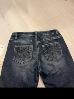 Blå jeans med broderade bakfickor - Snygga blå jeans med unika broderade detaljer på bakfickorna. Klassisk femficksmodell med normal passform och bälteshällor. Jeansen har en lätt tvättad look och är perfekta för dig som vill ha något extra i vardagen.