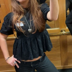 Svart blus med volang och struktur - Svart blus med volang nertill och puffiga korta ärmar. Blusen har en strukturerad yta och är croppad i modellen. Perfekt för dig som gillar en trendig och feminin stil. Jag brukar ha den bakåfram så knytningarna blir i ryggen men verkligen skitsnygg o ha de där fram me!😍
