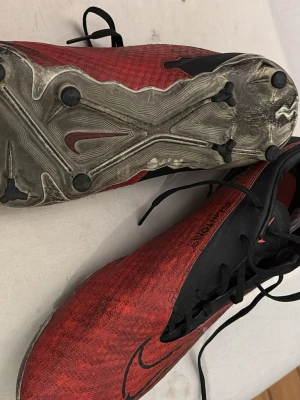 Röda Nike fotbollsskor med snörning - Snygga röda och svarta fotbollsskor från Nike med klassisk snörning och rund tå. Ovandelen är i syntetmaterial för bra bollkontroll och yttersulan har dobbar för optimalt grepp på planen. Perfekta för dig som vill ta ditt spel till nästa nivå.