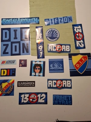 Djurgården klistermärken samling - En samling klistermärken med Djurgården-tema i olika former och storlekar. Färgerna är främst blå, gul och röd med motiv som DIF-logga, texter som 'Sofialäktaren', 'ACAB', 'DIFZON', 'FRIHET ULTRAS' och årtal. Perfekt för att dekorera prylar eller visa sitt stöd för laget.