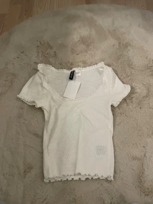 Vit ribbad topp med volangkant H&M - Söt vit ribbad topp från H&M med korta ärmar och volangkant vid ärmslut, halsringning och nederkant. Toppen har en djup rundringning och är tillverkad i mjuk bomullsmix. Perfekt för en enkel men gullig look.