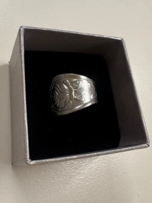 Bred silverfärgad ring med ros-motiv tillverkad på återvuuna bestick - Snygg bred ring i silverfärgad metall med ingraverat ros- och bladmotiv. Ringen har en bohemisk vibe och passar perfekt för dig som gillar unika accessoarer med lite vintagekänsla. 