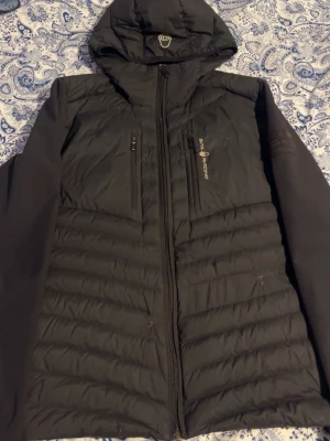 Svart dunjacka Sail Racing med huva - Svart dunjacka från Sail Racing med huva och två bröstfickor med dragkedja. Jackan har quiltad front och släta ärmar, samt logga på bröstet. Perfekt för kyliga dagar och har en sportig look. Finns lite skador på den som kan ses på bilderna mvh Robbin 