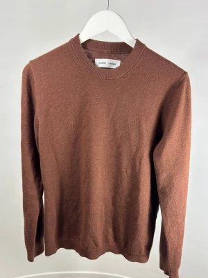 Samsøe Samsøe Stickad Tröja  - En enkel och stilren brunorange stickad tröja från Samsøe Samsøe med rund hals och långa ärmar. Modellen på bilden är 175 cm och väger 70 kg. Tröjan har ribbade muddar vid ärmslut och nederkant, vilket ger en klassisk look. Perfekt för lager på lager och passar till både jeans och chinos.