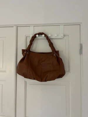 Brun handväska med flätat handtag - Säljer en snygg brun handväska i skinnimitation med ett unikt flätat handtag. Väskan är rymlig och sparsamt använd. Finns innerficka med dragkedja