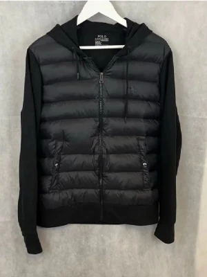 Svart cardigan från Polo Ralph Lauren - Svart cardigan jacka från Polo Ralph Lauren med huva och dragkedja framtill. Jackan har quiltad front i syntetmaterial, mjuka ärmar och två fickor med knappstängning. Perfekt för våren