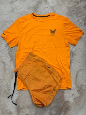 Nike Running Division - Snyggt träningsset från Nike med en orange t-shirt och matchande shorts. T-shirten har rund hals, kort ärm och tryckt logga med fjäril på bröstet. Shortsen har resår i midjan, dragsko och en liten ficka med dragkedja. Perfekt för löpning eller gym. Materialet känns lätt och andas. T-Shirt storlek: S Shorts storlek: L passar M