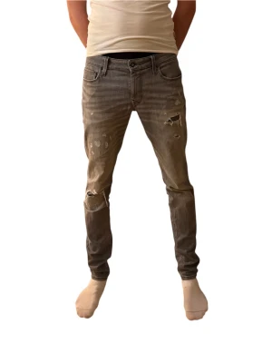 Grå slitna jeans från Jack & Jones - Säljer ett par grå jeans från Jack & Jones i storlek 33/36. Jeansen har skinny fit, slitningar och hål på benen för en riktigt trendig look. Klassisk femficksmodell med dragkedja och knapp. Perfekta för dig som gillar en edgy stil.