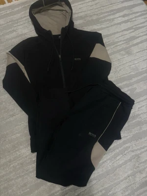 Svart och beige loungewear-set BOSS - Mysigt loungewear-set från BOSS i svart med beige detaljer. Setet består av en hoodie med dragkedja och matchande byxor, båda med diskret BOSS-logga. Perfekt för chill dagar hemma eller när du vill vara bekväm men ändå snygg.