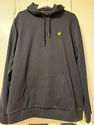 Svart hoodie från Lyle & Scott - Svart hoodie från Lyle & Scott med klassisk gul logga på bröstet. Hoodien har huva med dragsko och en stor magficka framtill. Perfekt för en chill och stilren look. Endast testad