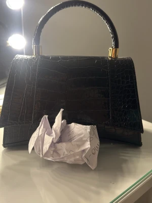 Svart handväska i krokopräglat skinn - Stilren svart handväska med krokodilpräglat mönster och guldfärgade detaljer på handtaget. Väskan har en klassisk, strukturerad form och ett stadigt handtag. Perfekt accessoar för att lyfta din outfit. Guldigt b vid pappersbiten#FAK€