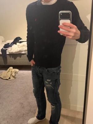 Svart kabelstickad tröja Polo Ralph Lauren - Kabelstickad svart tröja från Polo Ralph Lauren med rund halsringning och röd broderad logga på bröstet. Tröjan har långa ärmar och ribbade muddar vid ärmslut och nederkant. Perfekt för lager-på-lager och snygg till jeans.