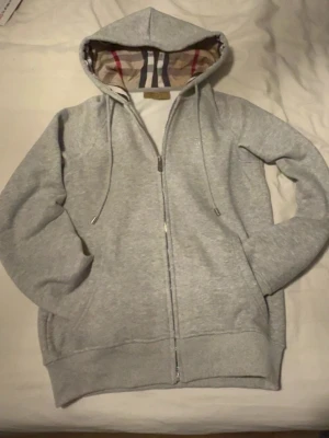 Grå zip hoodie från Burberry Dam - Snygg grå zip hoodie från Burberry med klassiskt rutigt foder i huvan och dragsko. Tillverkad i mjukt bomullsmaterial och har två fickor framtill. Perfekt för dig som vill ha en stilren och exklusiv hoodie med ikonisk Burberry-touch. Storleken är M men är mer som S. Använd en gång.